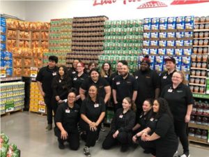 East Tacoma Grocery Outlet employees pose in their uniforms in front of grocery display and mural of Mount Rainier and Tacoma Dome.