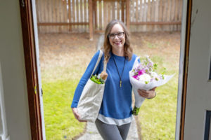 Image of door open to Give InKind founder Laura Malcom, carrying a bouquet and bag of groceries.
