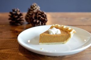 Image of pumpkin pie slice on plate with pine cones and tabletop in background