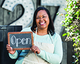 Image of mature, African American woman wearing an apron is standing outdoors holding an OPEN sign.